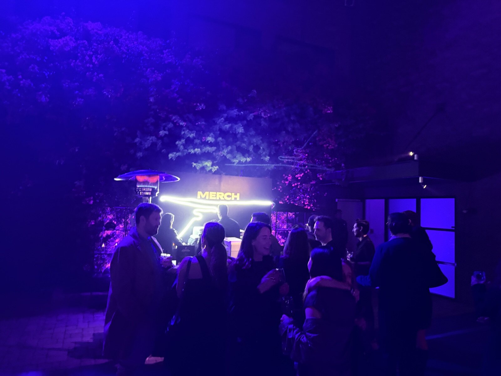 Glowing MERCH sign above an outdoor night-time merch bar with a crowd of guests under blue and purple light