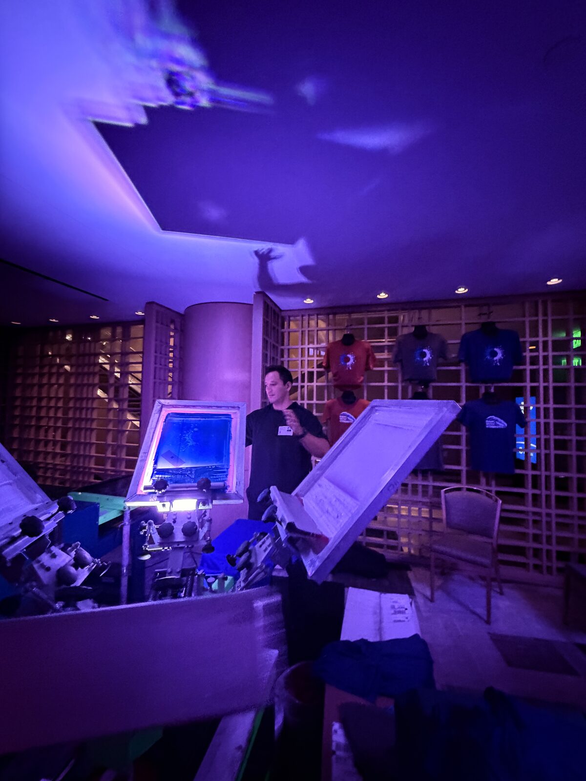 Crew member at a screen printing press under UV light with printed tees glowing on a lattice wall behind him
