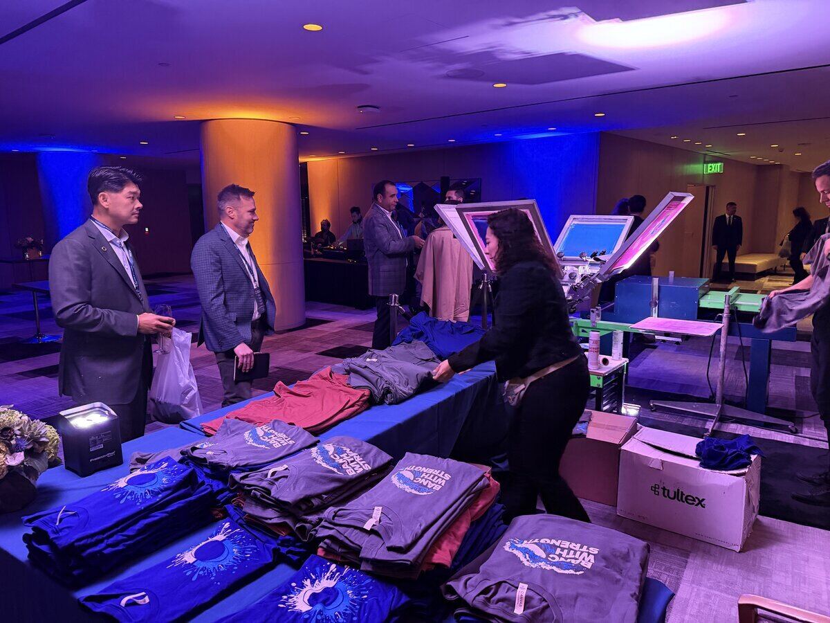 Folded event tees in three colorways staged on a long blue table beside two screen printing presses in a ballroom