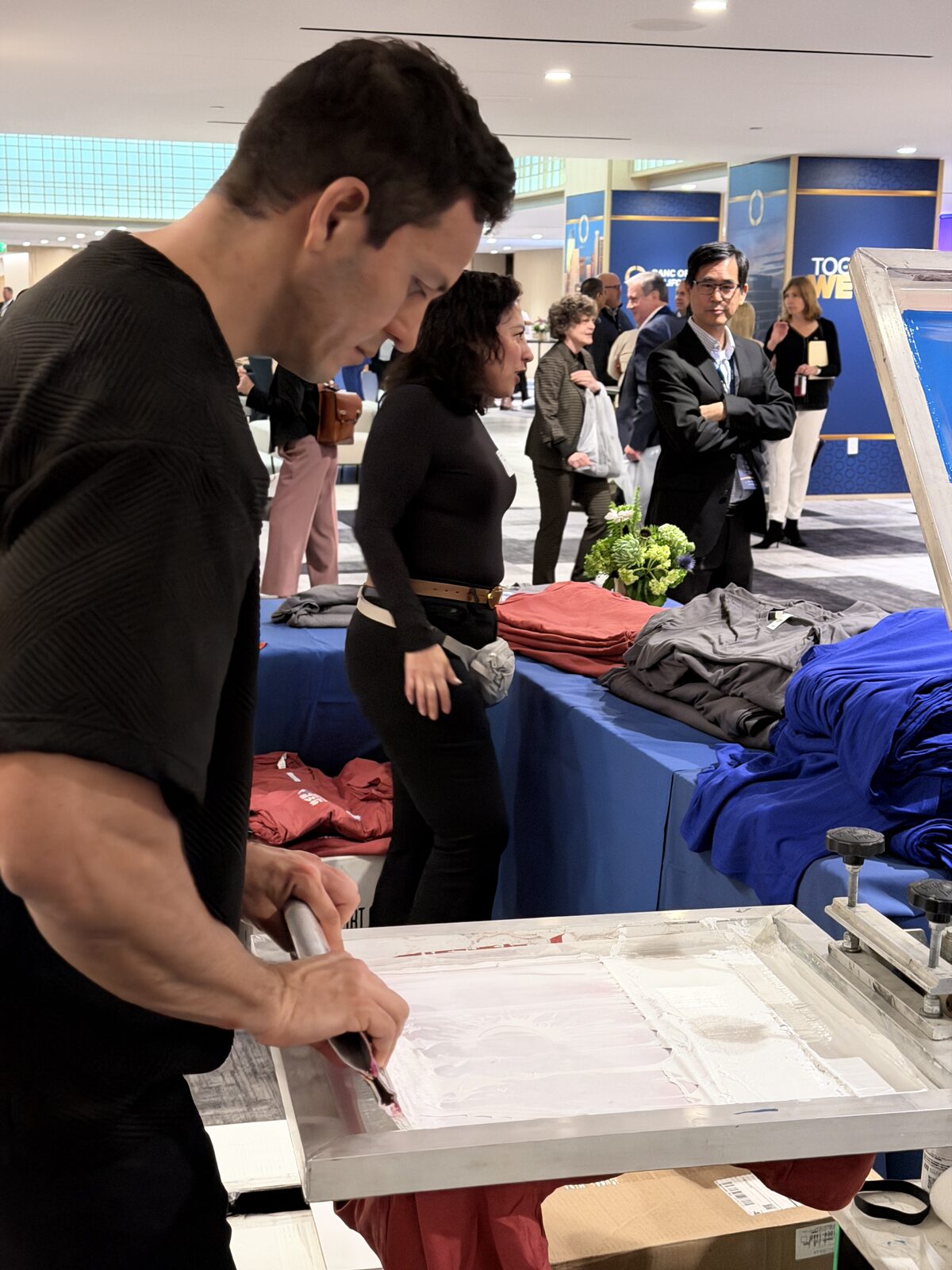 Conference guests pausing to watch a screen printer flood a screen with white ink beside a table of folded shirts