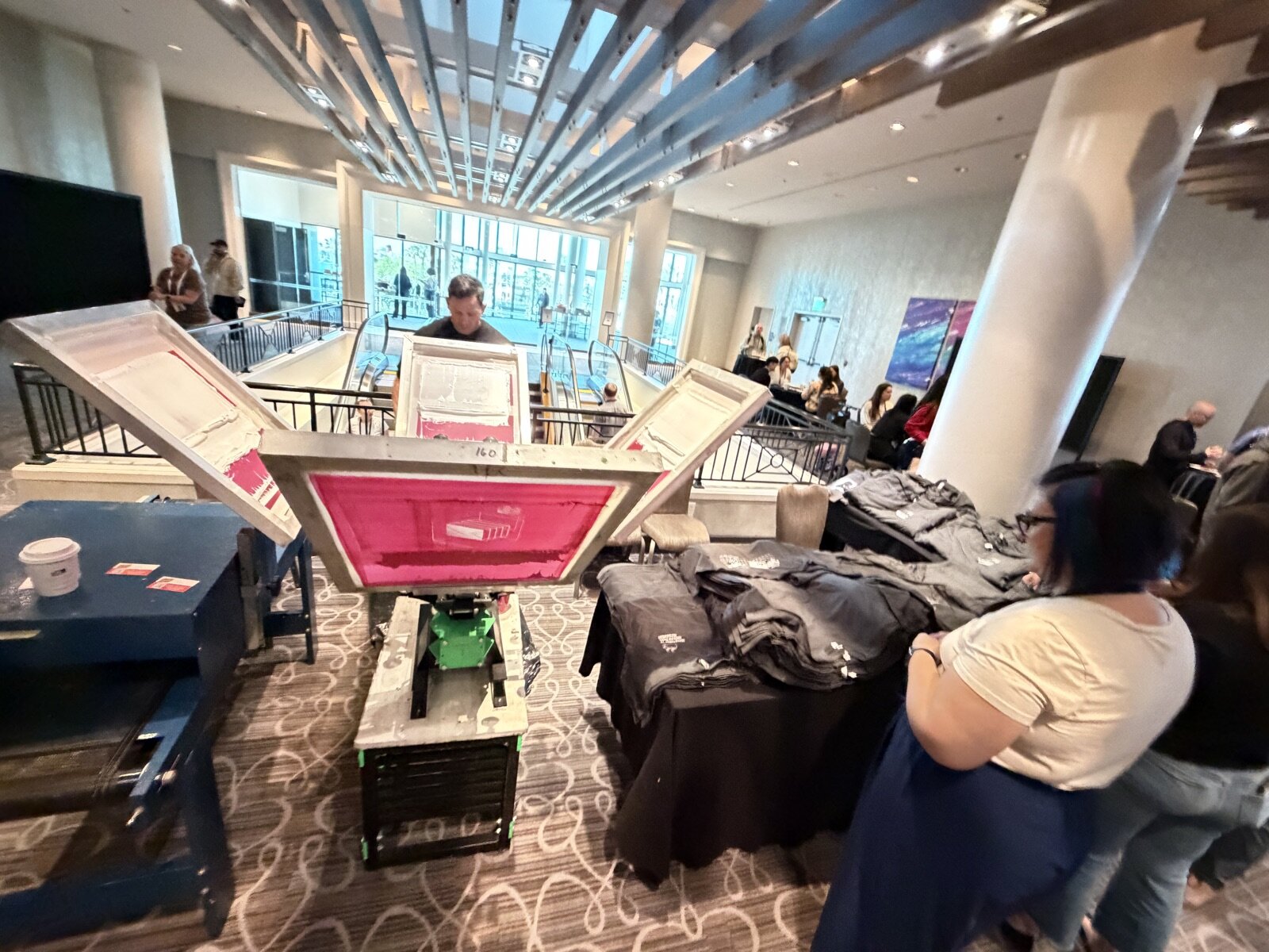 Manual screen printing press with open screens beside stacks of folded tees in a hotel conference foyer