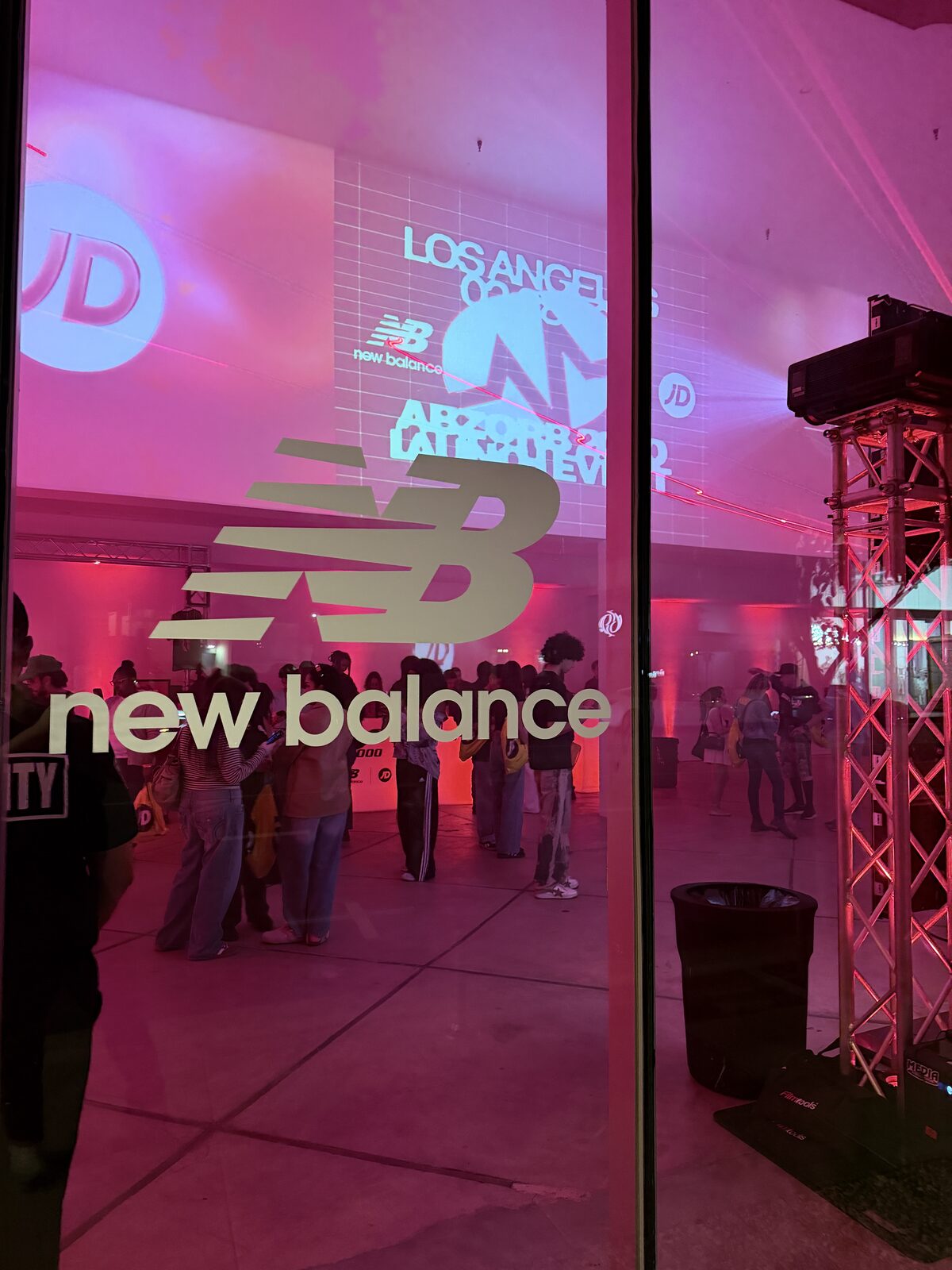 Gold New Balance logo on a window with launch party guests gathered inside a pink-lit venue